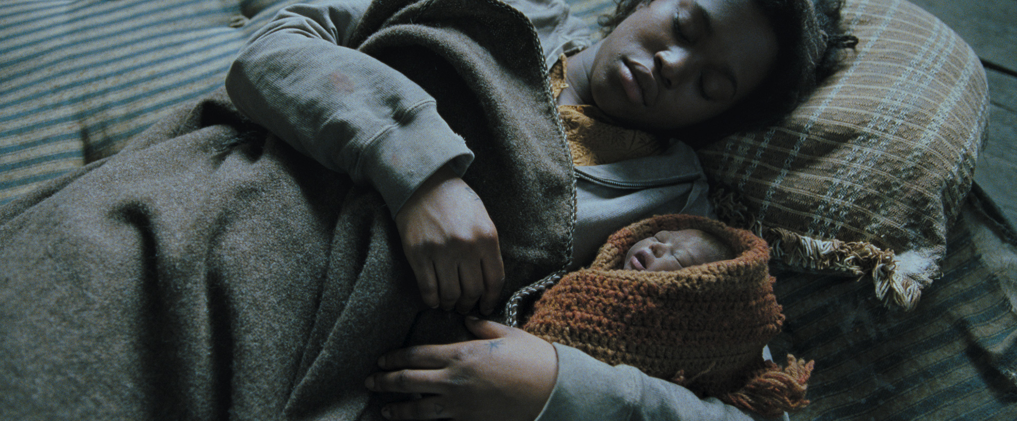 A young black woman lies asleep, under a coat, holding a newborn baby