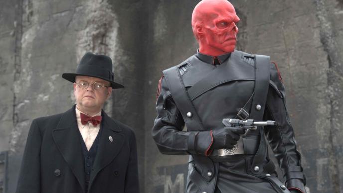 doctor arnim zola and red skull from captain america the first avengers standing next to each other. doctor arnim zola is wearing a suit, bow tie and fedora as he looks directly into the camera and red skull is wearing a leather suit holding a gun and looking to the right