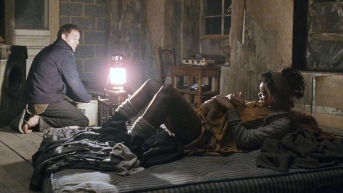 actor clive owen and actress julianne moore from children of men in an abandoned room. julianne moore is laying on a mattress holding a newborn baby as clive owens washes clothes in a bucket