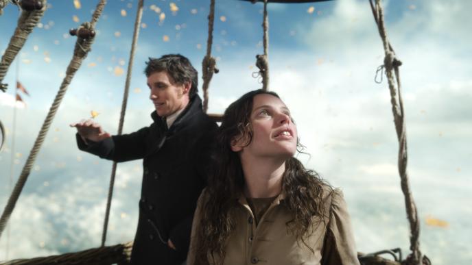actors eddie redmayne and felicity jones from the aeronauts standing in a hot air balloon in the sky surrounded by clouds and butterflies