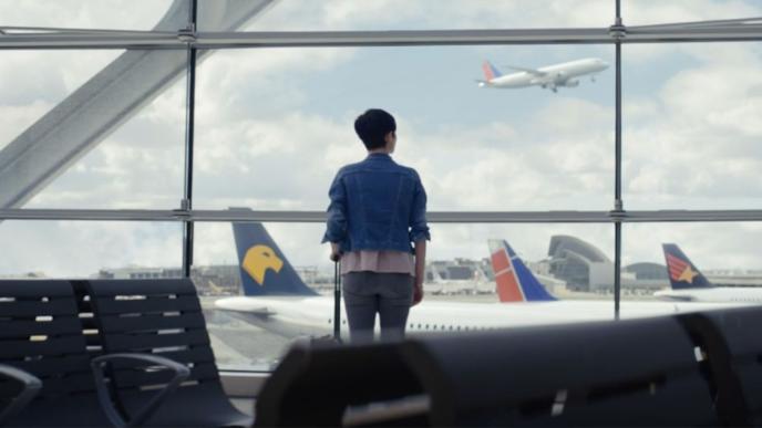 A woman stands with her back to us looking out of the window at an airport. There is a plane taking off in the background.