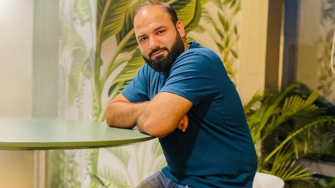 A man wearing a blue T-shirt sitting at a table, there is a wall behind him with foliage wallpaper. 