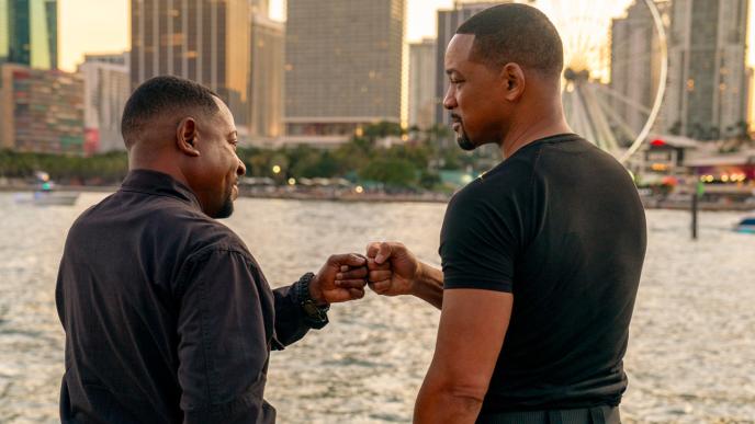 Two black men fist bumping in front of a body of water and a city skyline. 