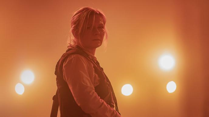 A woman wearing a bullet proof vest stood in the middle of a road with bright spotlights behind her. The air is filled with dust and has an orange hue to it.