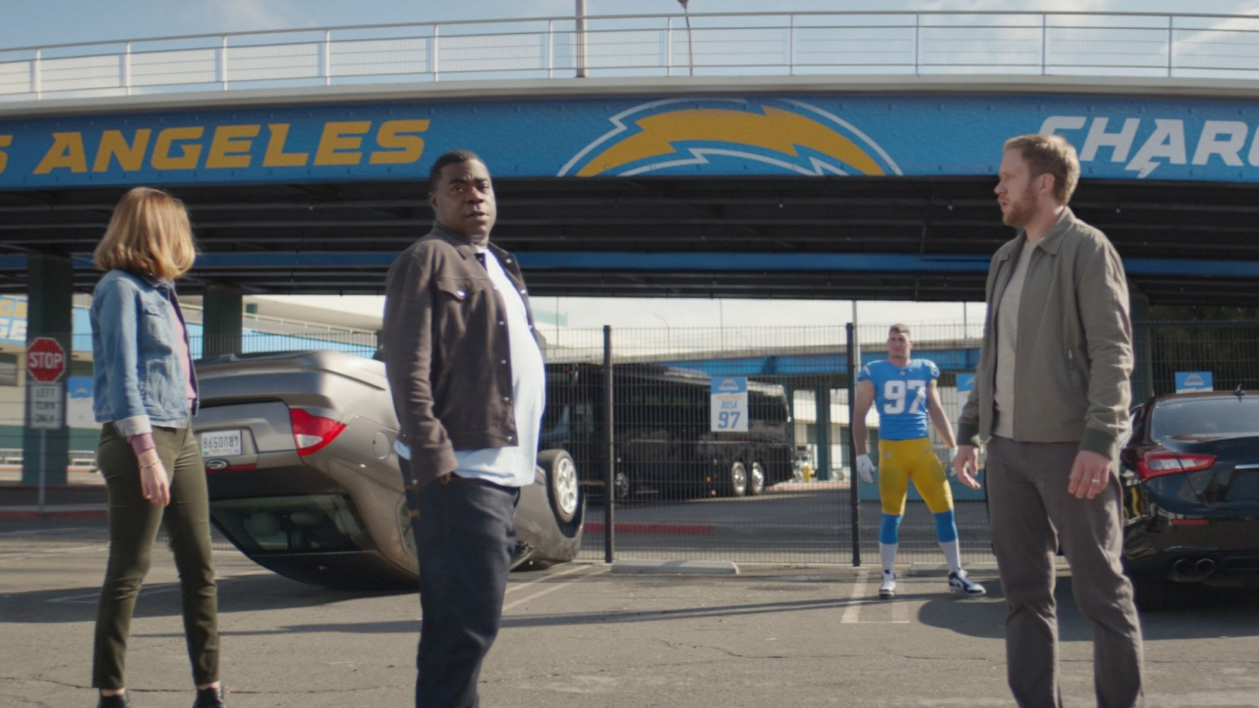 three people standing in front of a flipped car. there is an american football player in the background