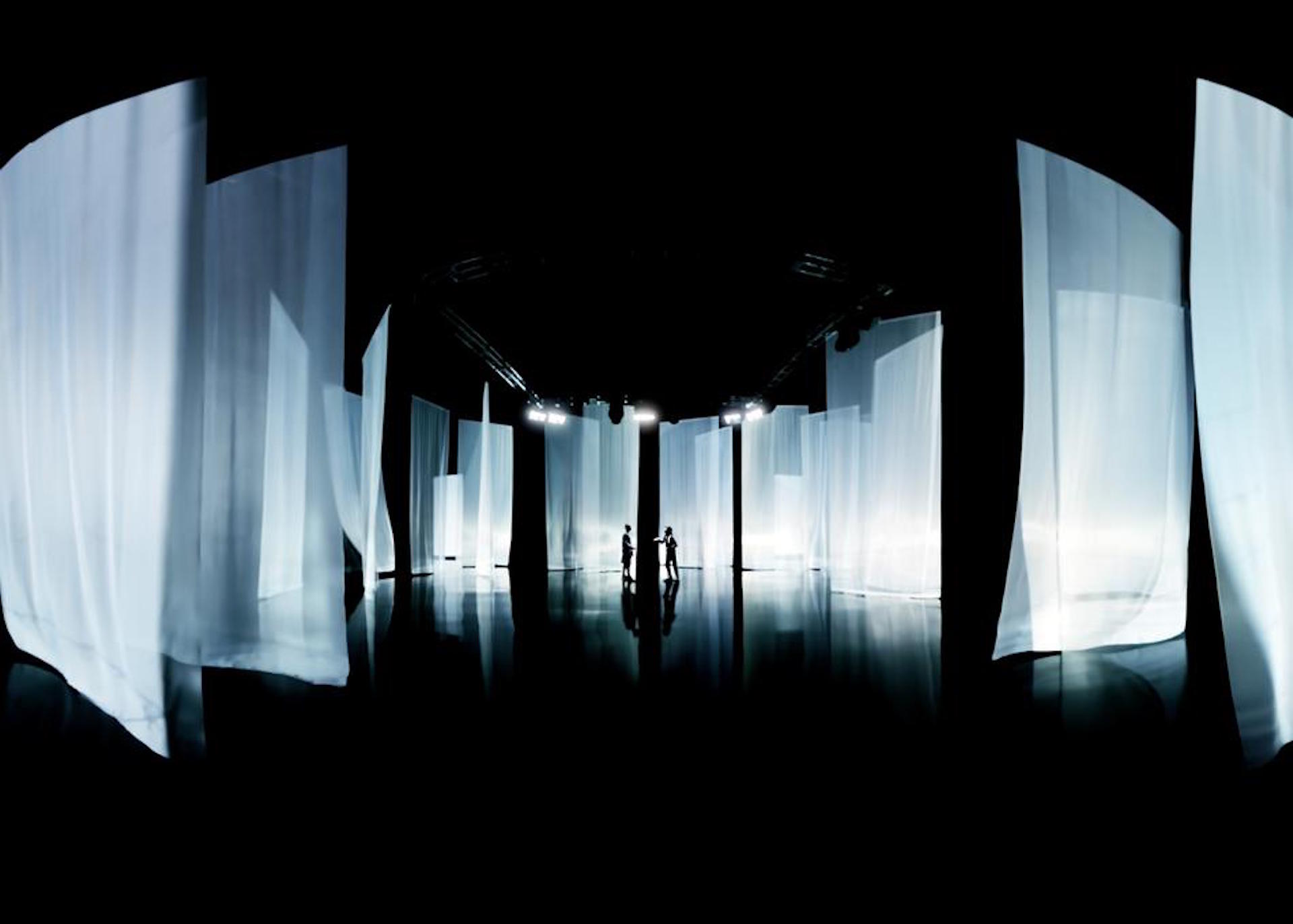 two people standing in the centre of an exhibition that has full length sheer sheets hanging from the ceiling