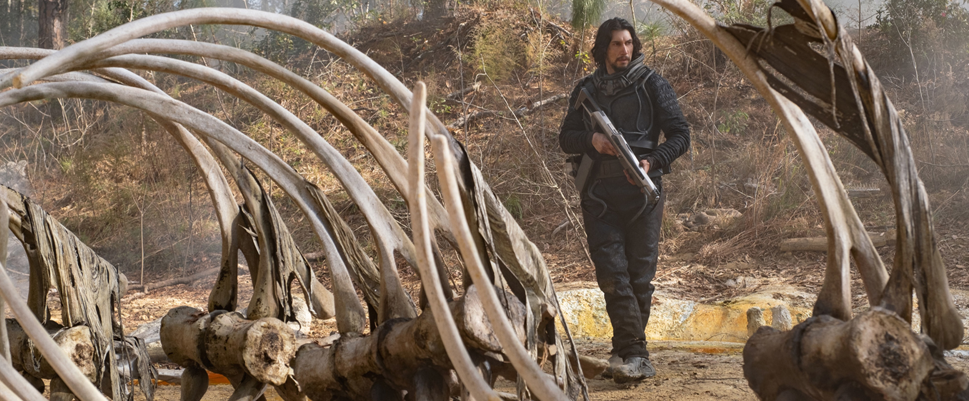 Adam Driver holds a large gun, standing near a large skeleton