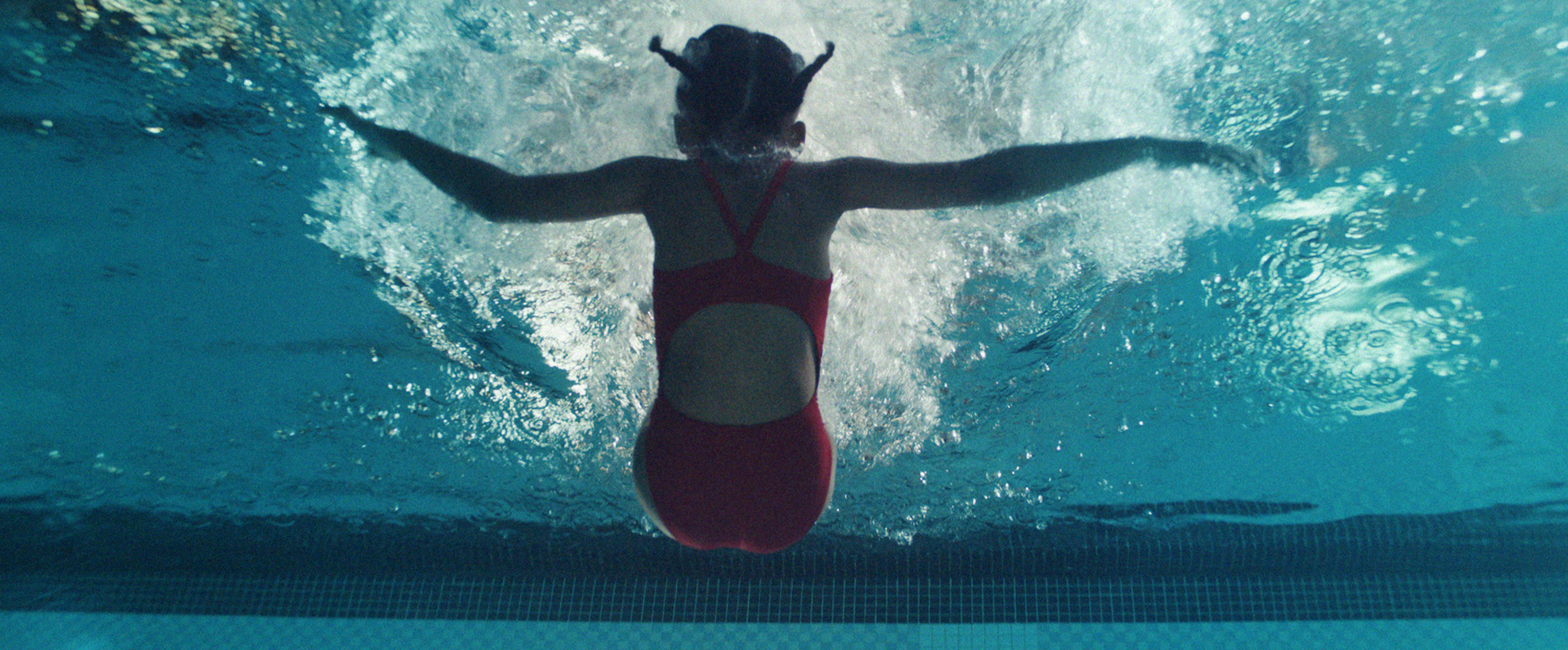 A young girl in a red swimming costume falls backwards into a pool. The shot is from the bottom of the pool, looking upwards.