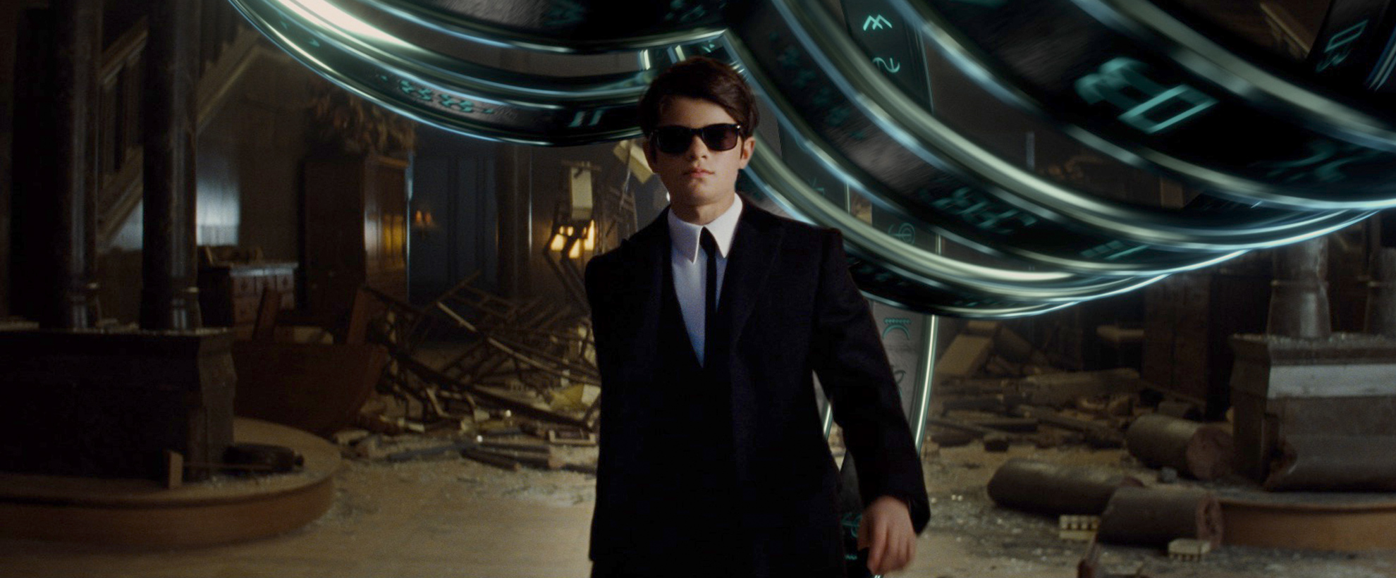 A young boy in a black suit and sunglasses walks towards the camera