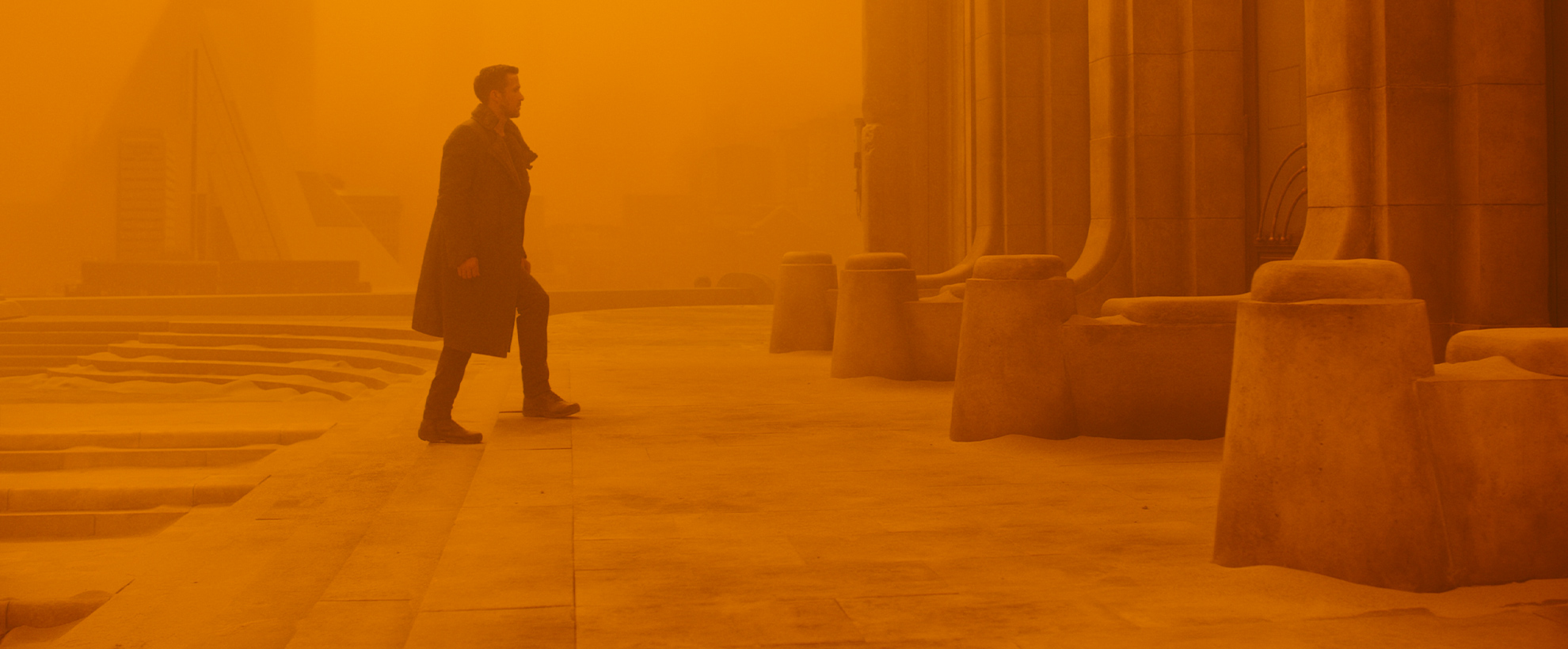Ryan Gosling walks up steps towards stone pillars, the whole image is saturated in orange tones