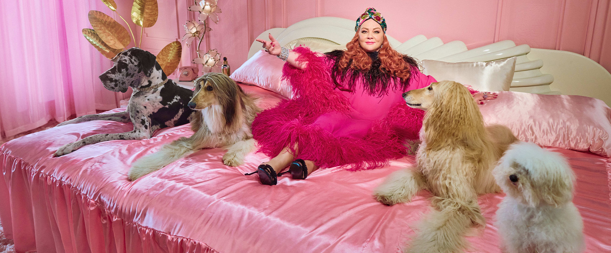 Melissa McCarthy sits on a pink satin bed, wearing a flamboyant pink robe, surrounded by different dogs