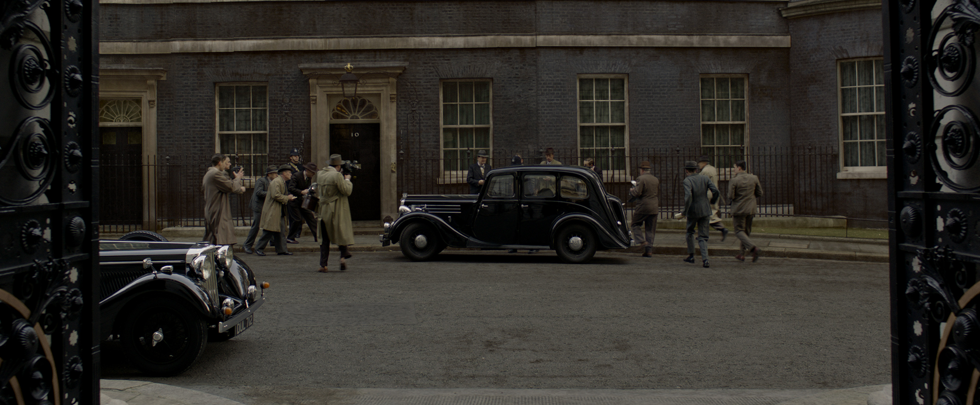 A vintage black car pulls up outside number 10 Downing Street, pursued by men on foot