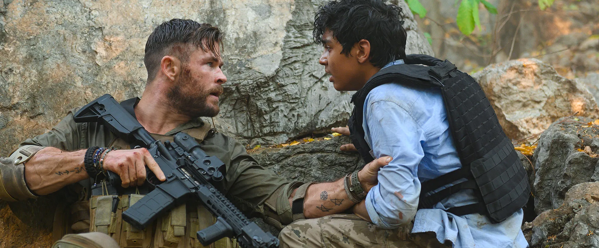 Chris Hemsworth crouches against a wall, dressed in military gear, holding the arm of a young boy in a bulletproof vest