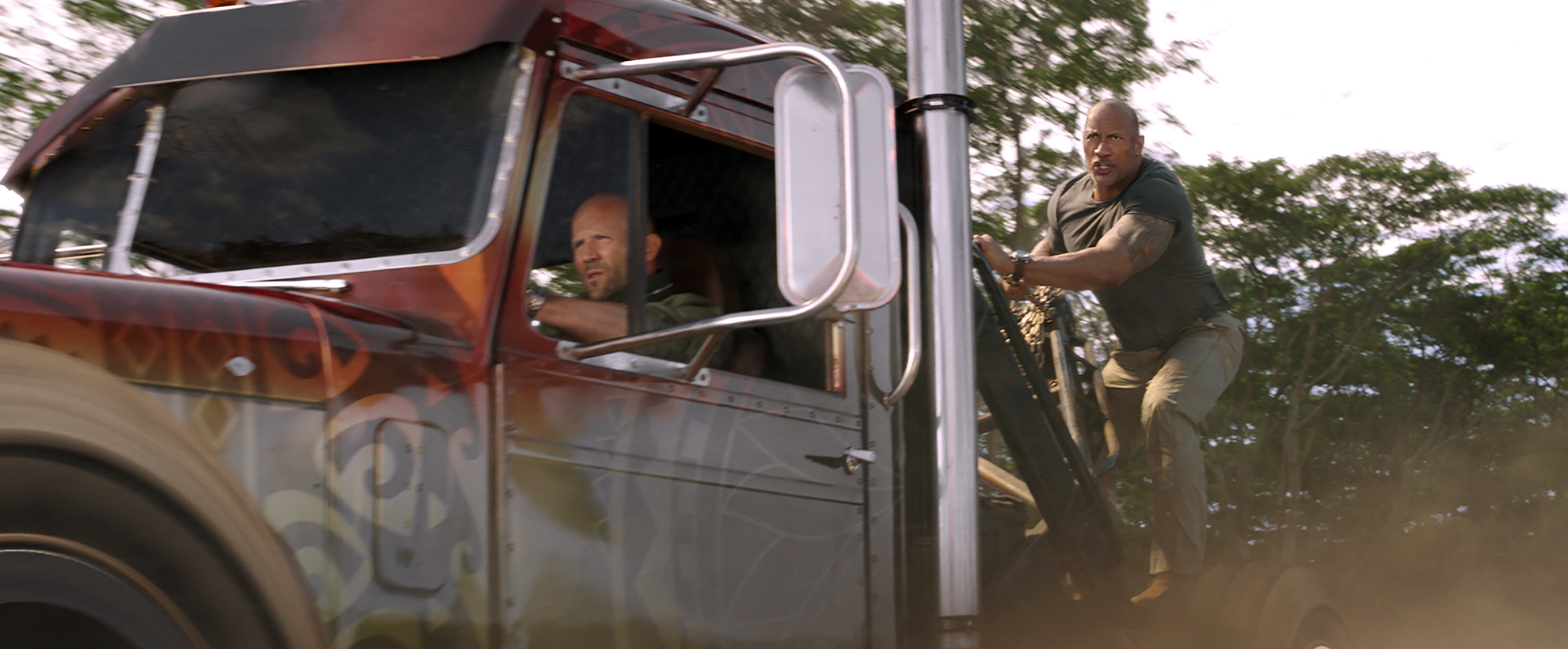 Jason Statham driving a large truck with Dwayne Johnson hanging on to the outside