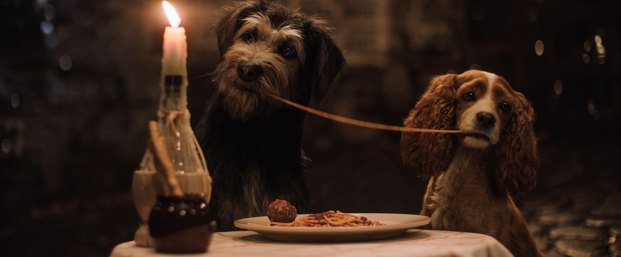 Two dogs sit at a candlelit table, eating either end of one spaghetti strand
