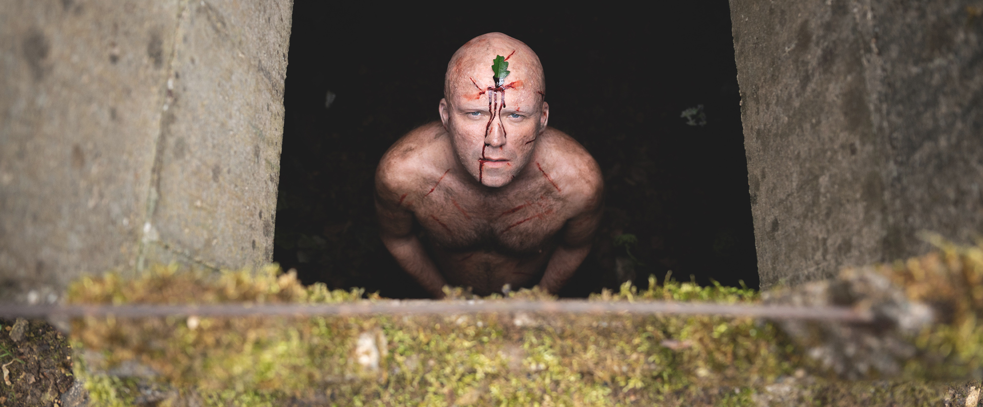 A nude man stands in an underground tunnel, with a leaf in his forehead