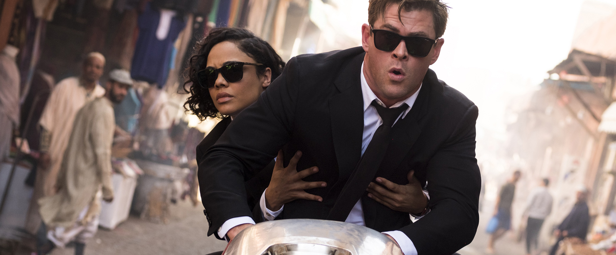 Chris Hemsworth and Tessa Thompson ride a motorbike, wearing black suits and sunglasses