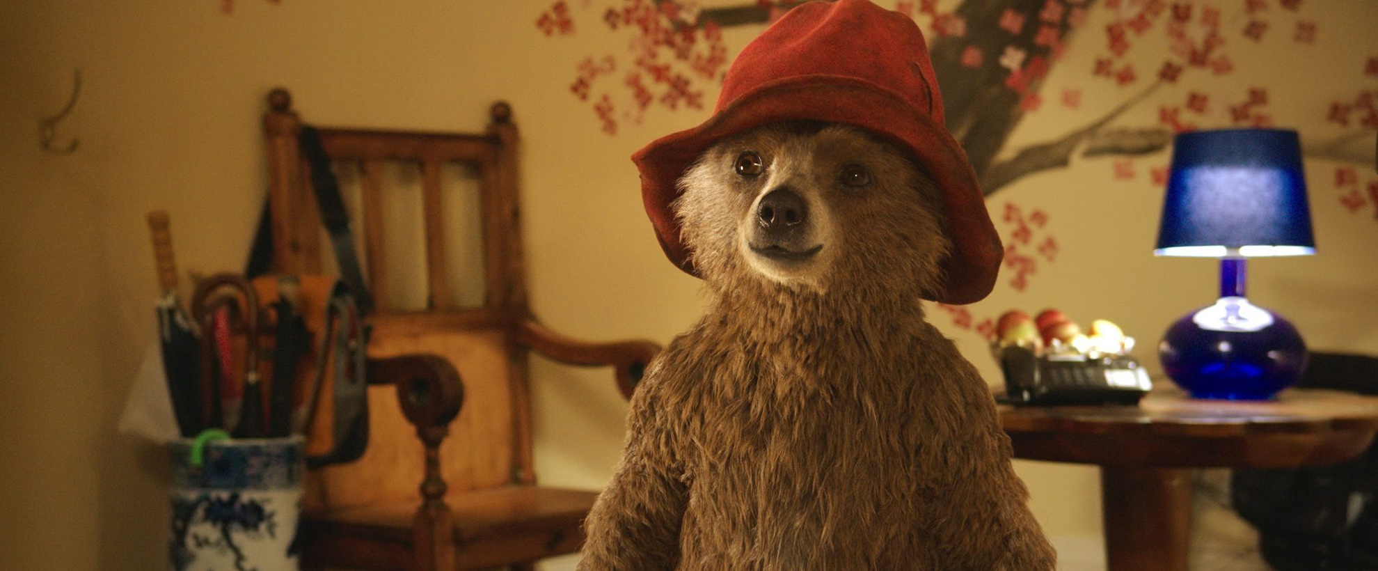 Paddington stands, wearing a red hat, in the hallway of the Brown's home