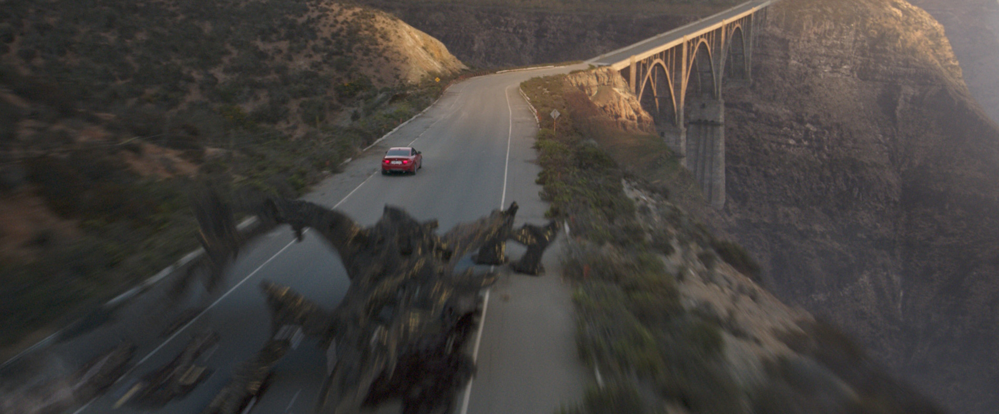 A red car drives down a mountain road at sunset, chased by an alien creature