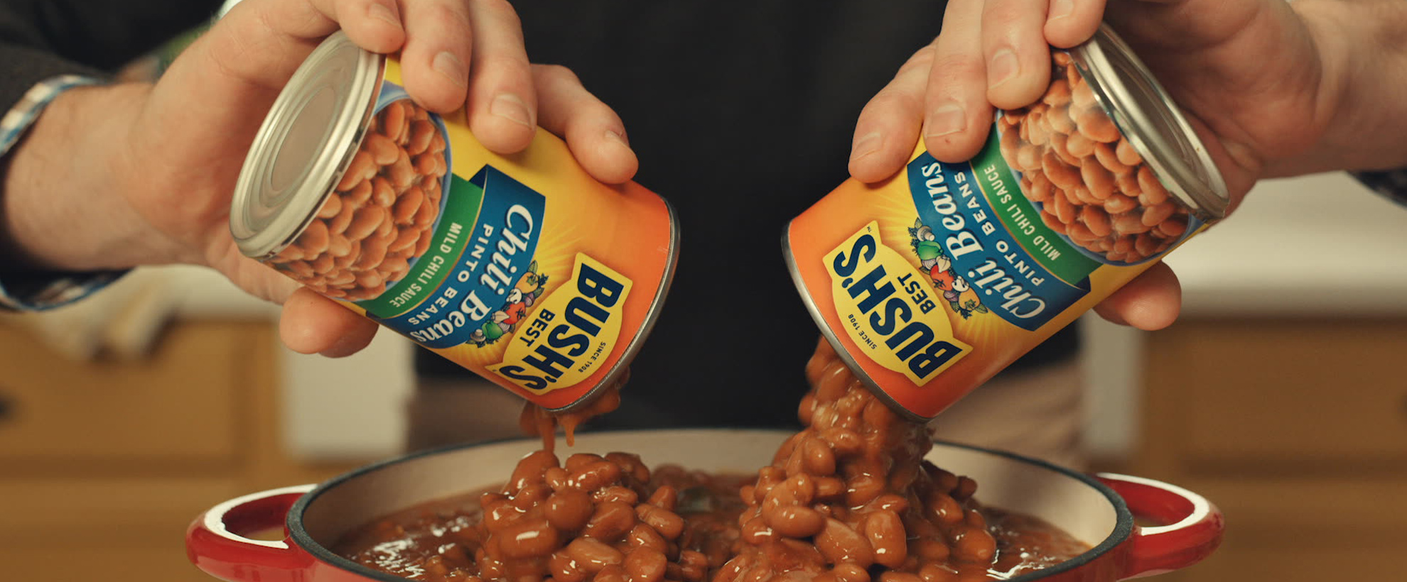 A man holds a can of Bushs beans in each hand, pouring them onto a plate