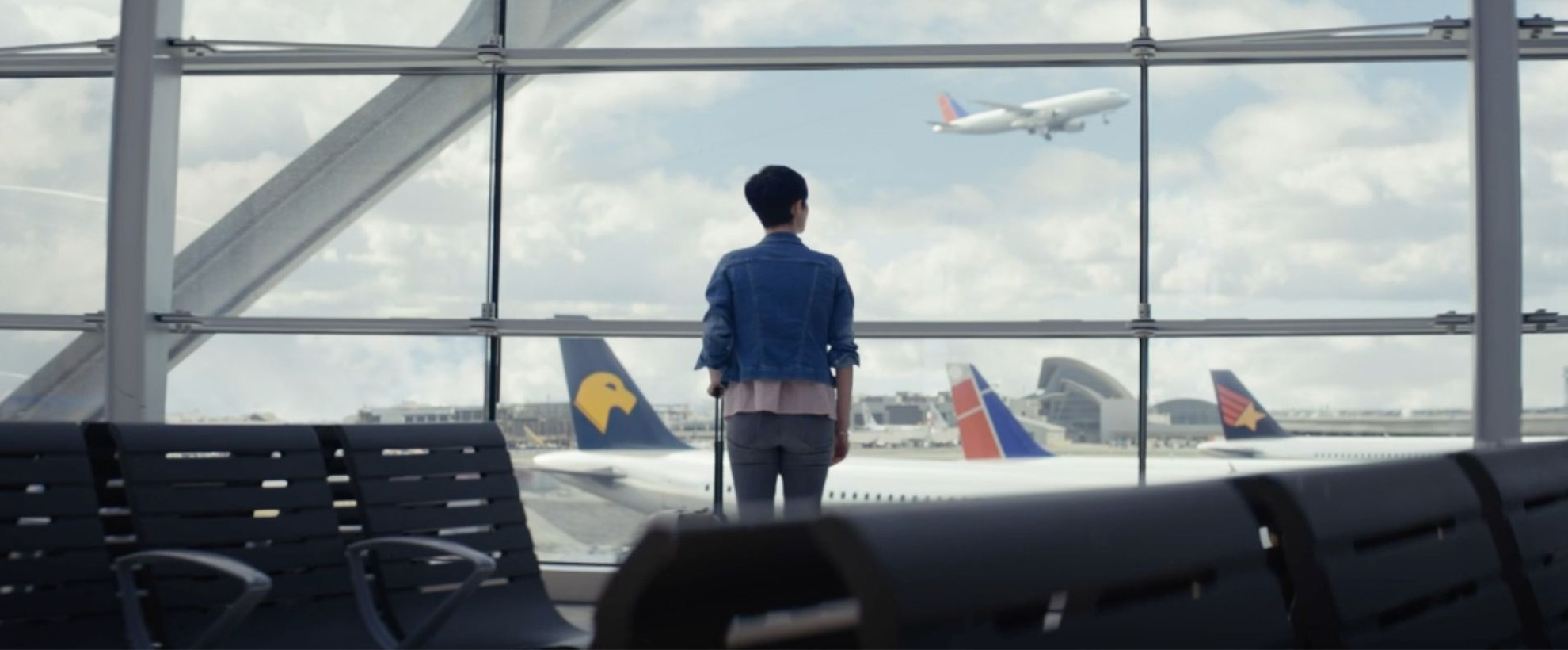 A woman stands with her back to us looking out of the window at an airport. There is a plane taking off in the background.