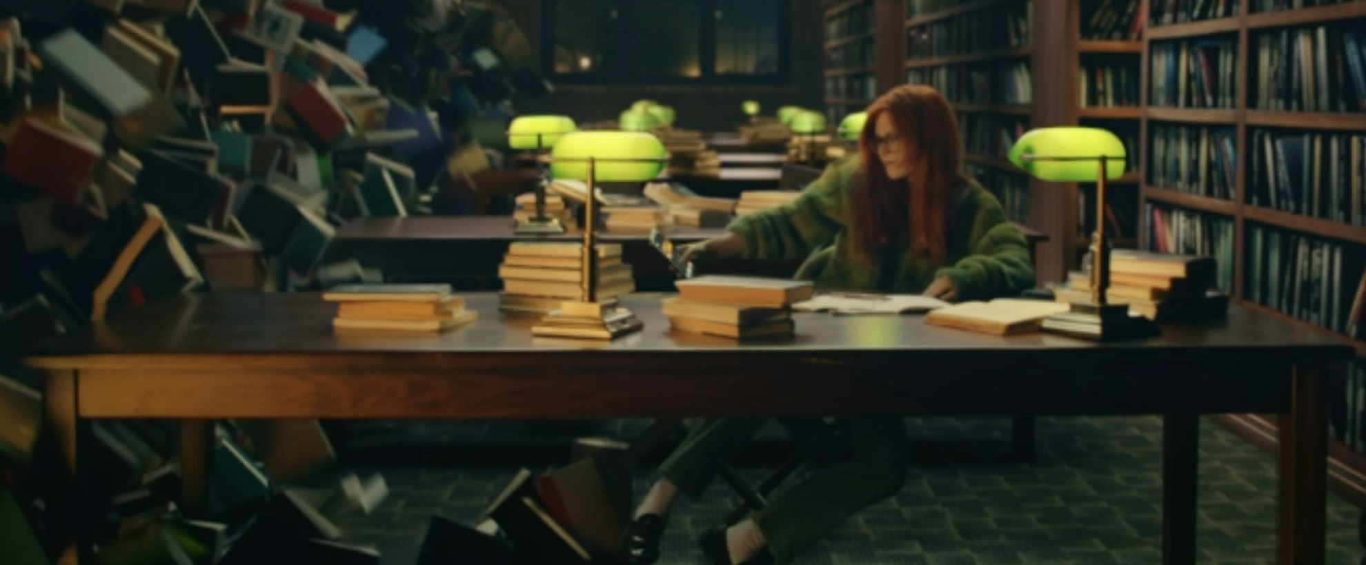 A woman in a green jumper sitting at a desk in a library. There are books falling down from the shelves towards her.