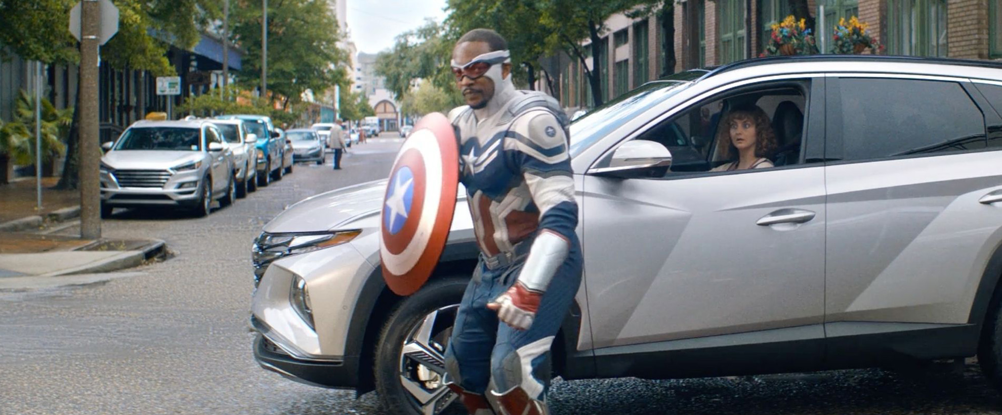 Captain America holding his shield standing in front of a silver car in the middle of the street.