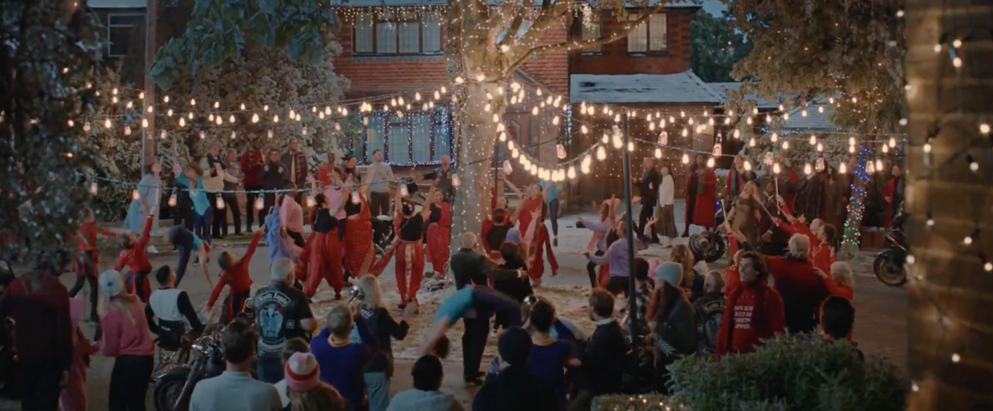 A large group of people dressed in red and holiday clothing celebrate outside under Christmas lights