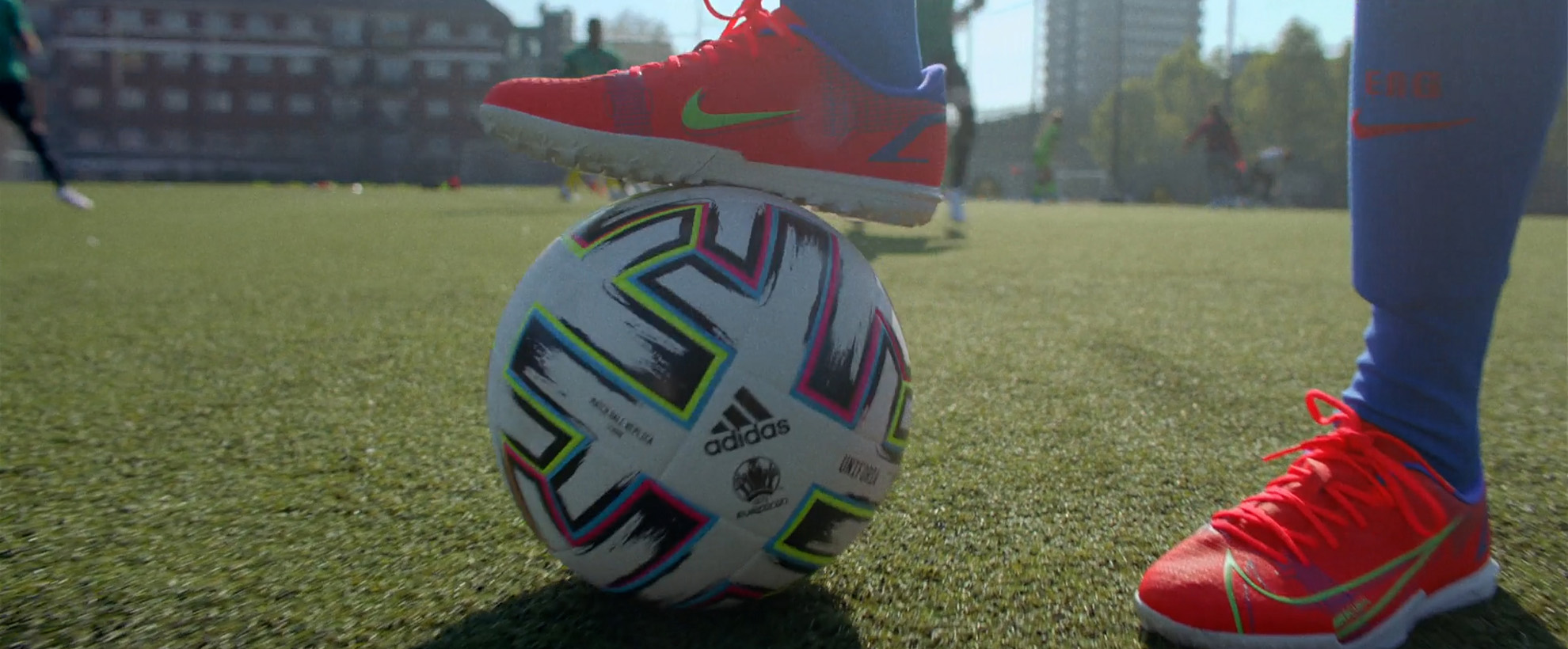 A pair of feet in orange trainers, one of the feet is on top of a football on a football pitch.