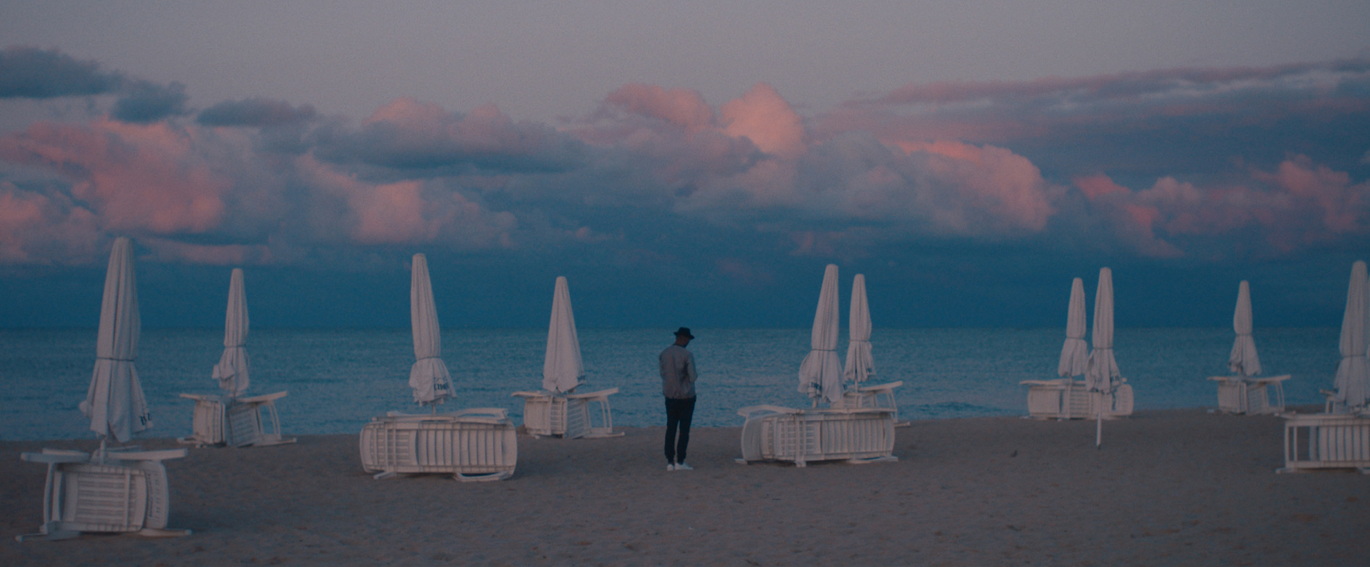 a person standing on the beach looking at the sea and mountains that are blankeed with pinky clouds. there are closed umbrellas and tables dotted around the beach