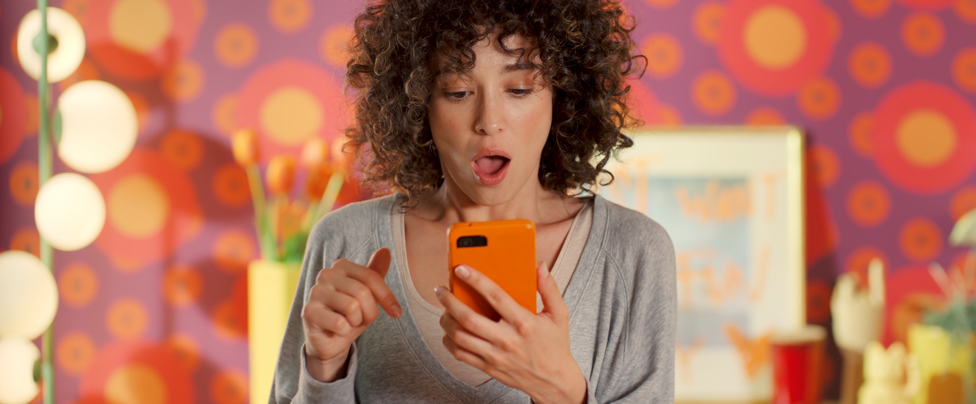 A young woman stares at a mobile phone with an orange cover, in a brightly coloured bedroom