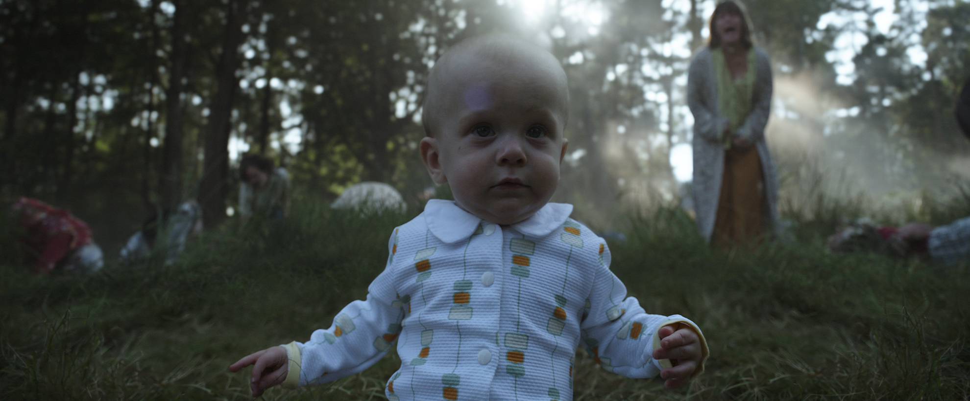 A baby toddles towards the camera, in a forest setting, with a woman screaming in the background, out of focus