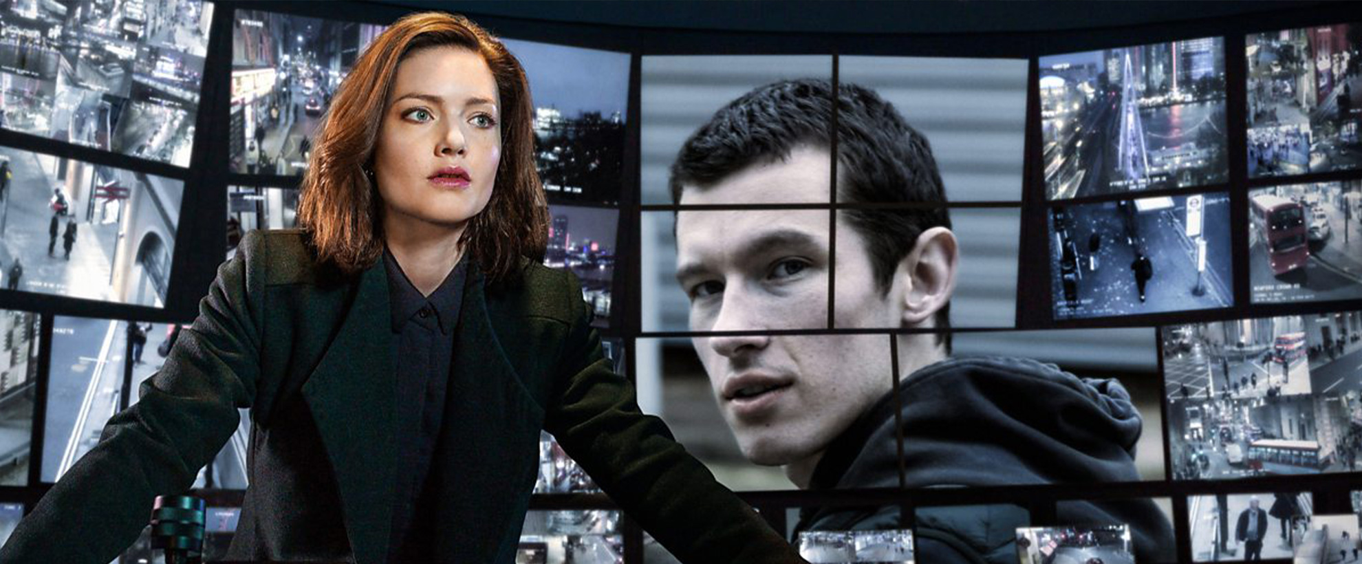 A red haired woman in a smart black suit, stands at a desk surrounded by screens