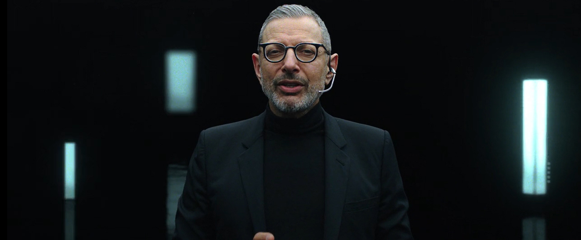 Actor Jeff Goldblum stands in a dark room. He is wearing a black blazer, black turtleneck and black thick framed glasses. He is also wearing a head piece