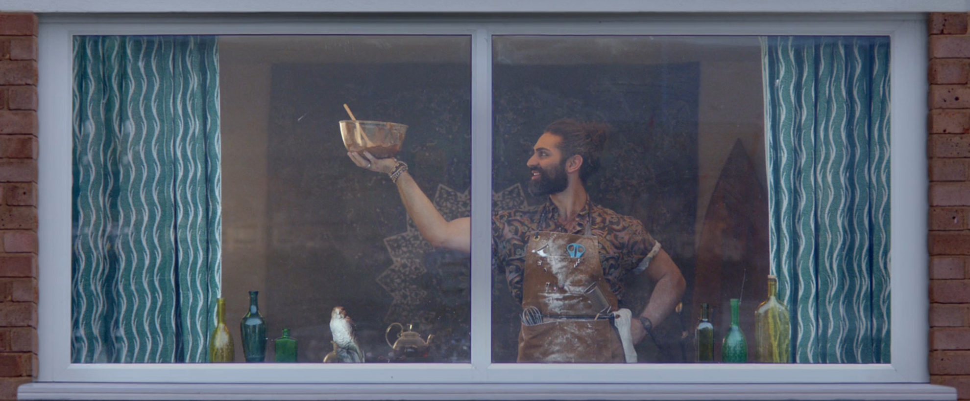 A man is seen through a window. He wears a work apron and proudly holds a bowl of batter