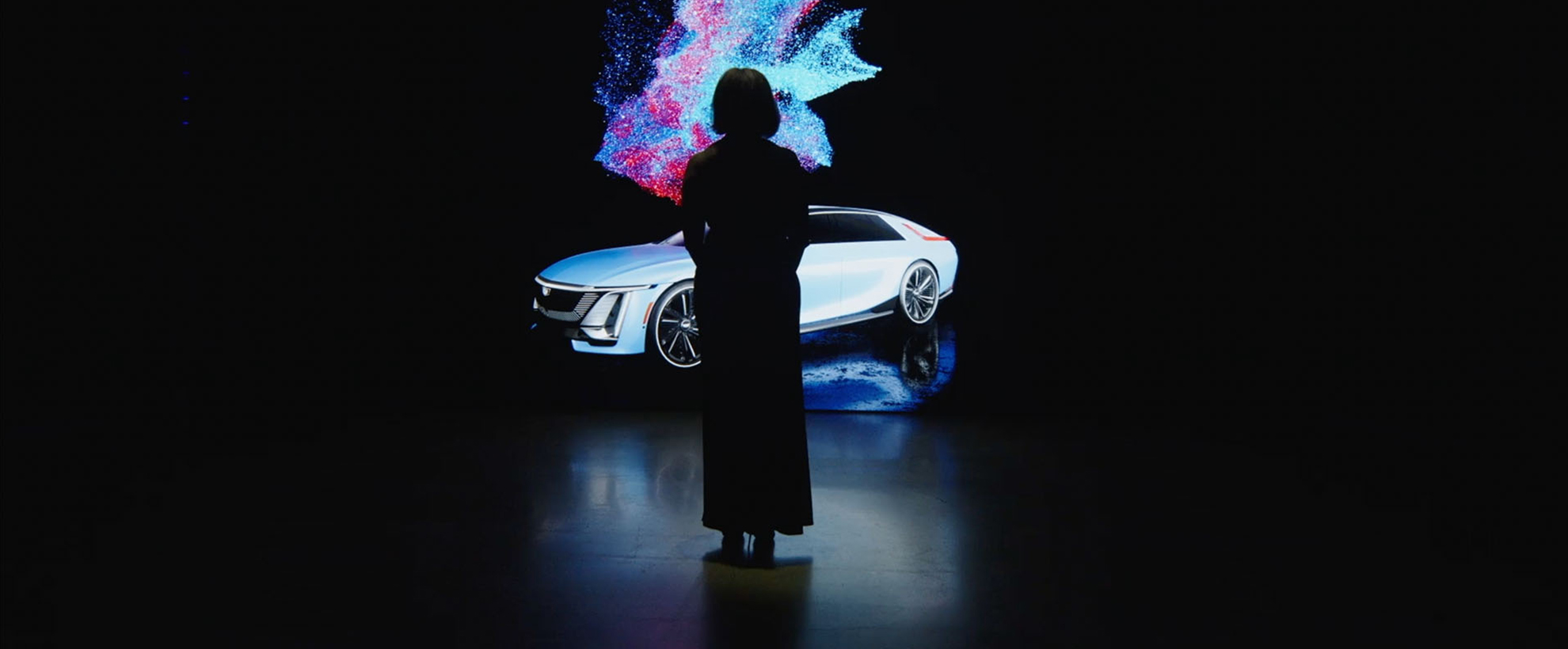 The silhouette of a person as they stand in front of a large screen in a dark room. On the screen is the Cadillac Lyriq car