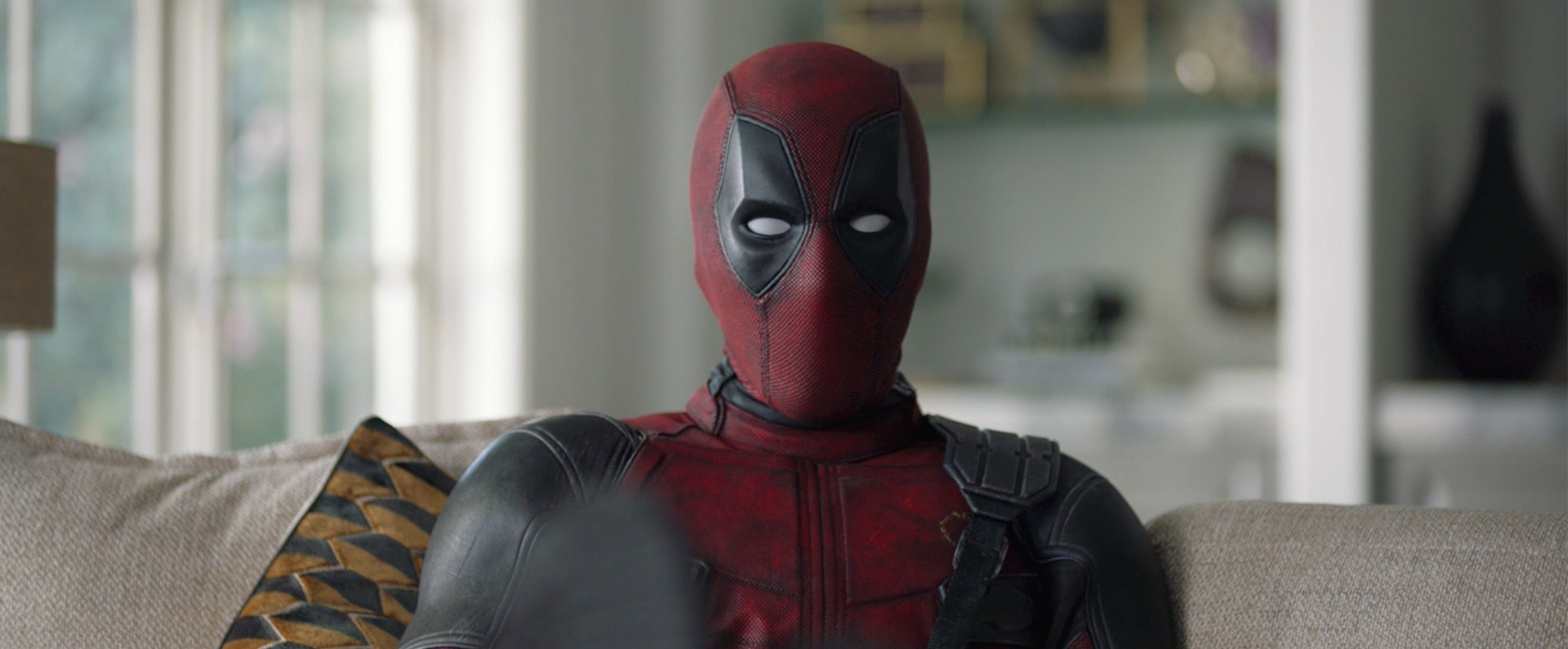 Deadpool is sat on a sofa looking at the camera with his feet up.