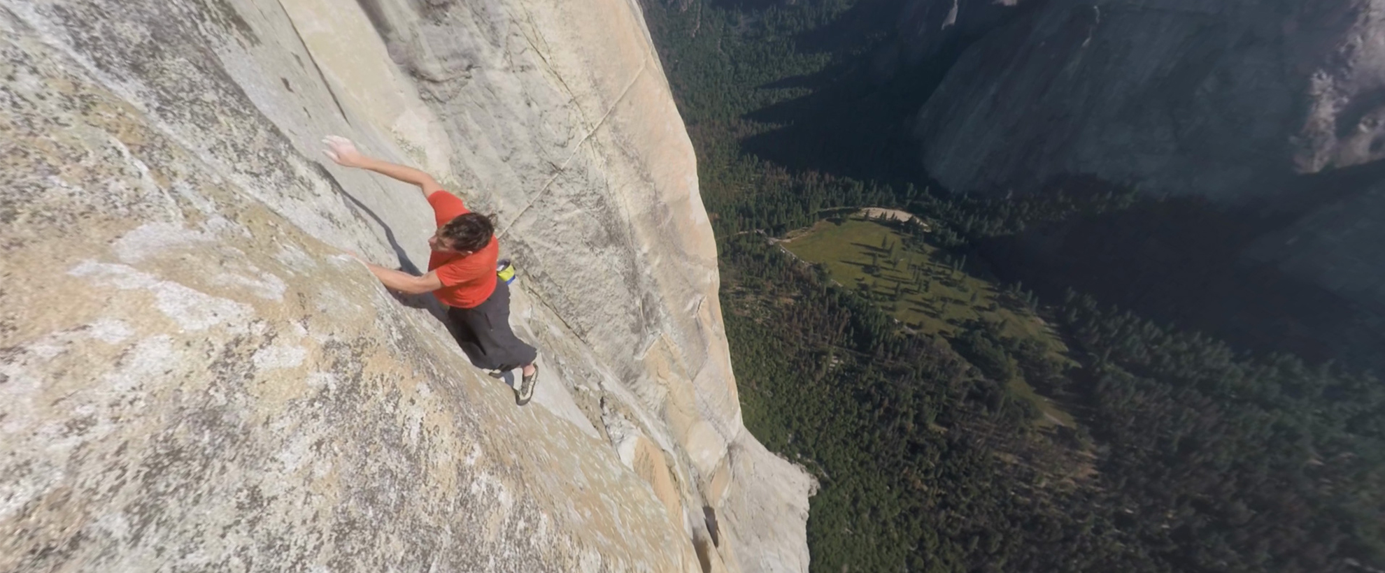 A man free climbing a large rock face above a forested hillside.