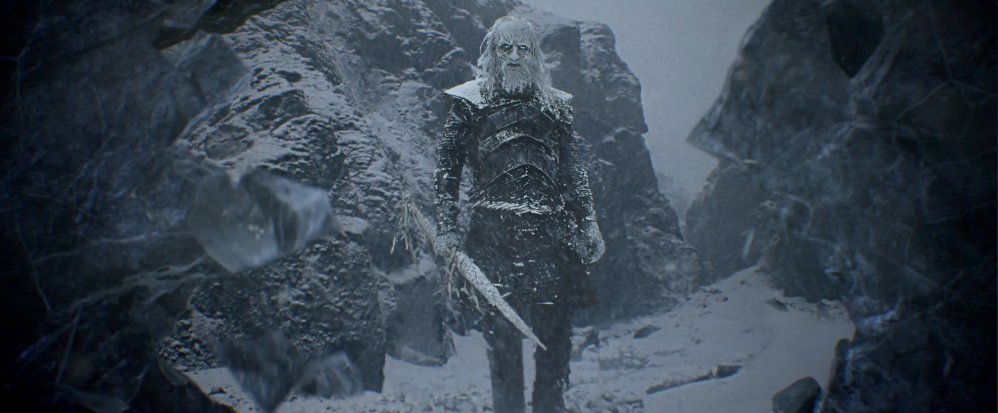 White Walker from Game of Thrones stands in a snowy rocky terrain. He is pure white with ice blue eyes, wears gray armor and holds an icy sword