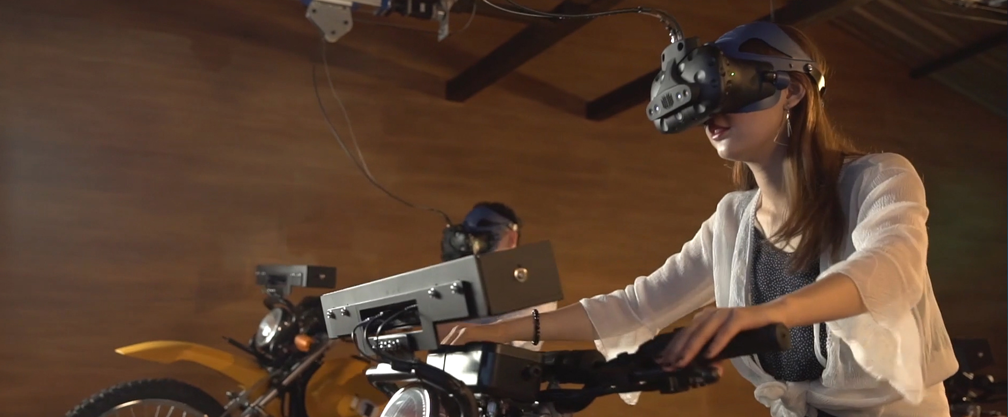 A woman wearing a white shirt sits on a motorcycle indoors with a VR headseat on her head