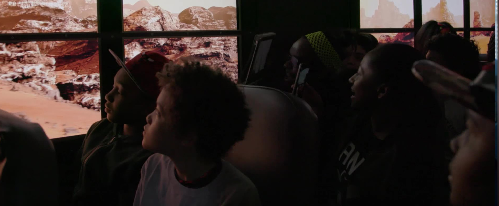 A group of school children in a school bus look out the bus windows into what looks like the planet Mars. The windows are replaced by screens which create the effect that the bus is driving on Mars