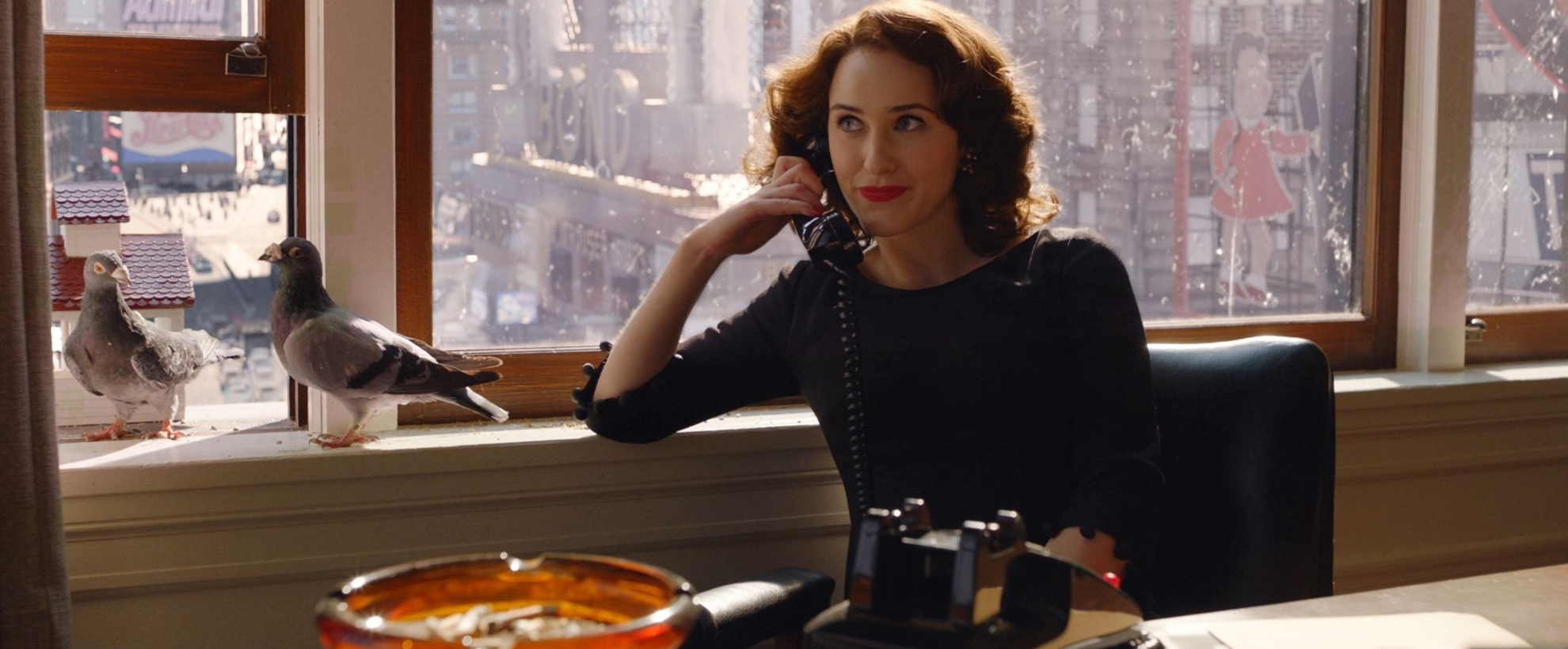 Miriam 'Midge' Maisel sits at a desk chair with a cityscape behind her seen through the windows