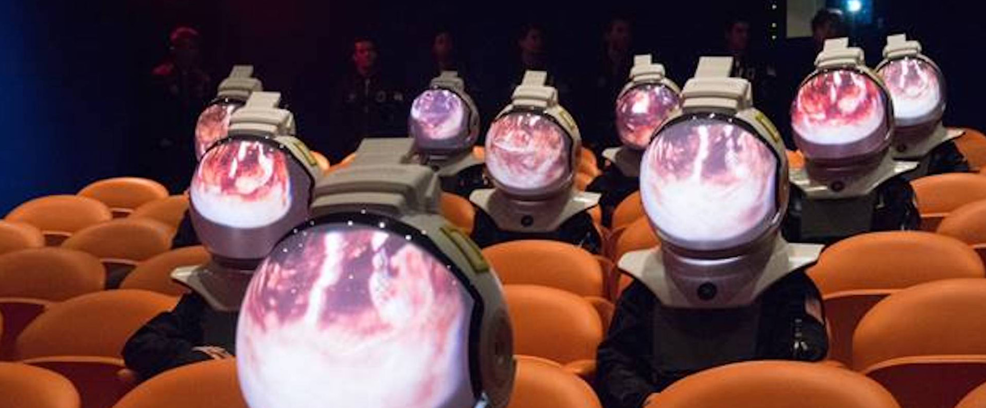 A group of people in space suits sit in orange chairs. In the reflection of their space helmets is a planet