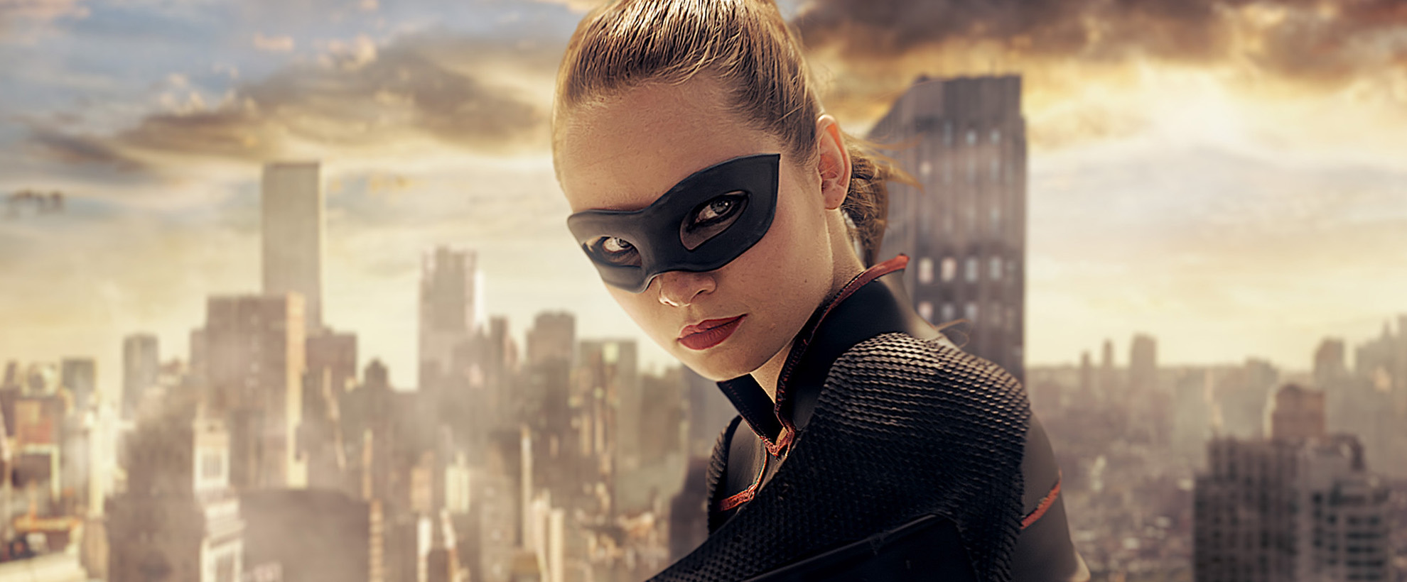 A woman with a superhero mask  standing on the top of a building with a cityscape in the background.