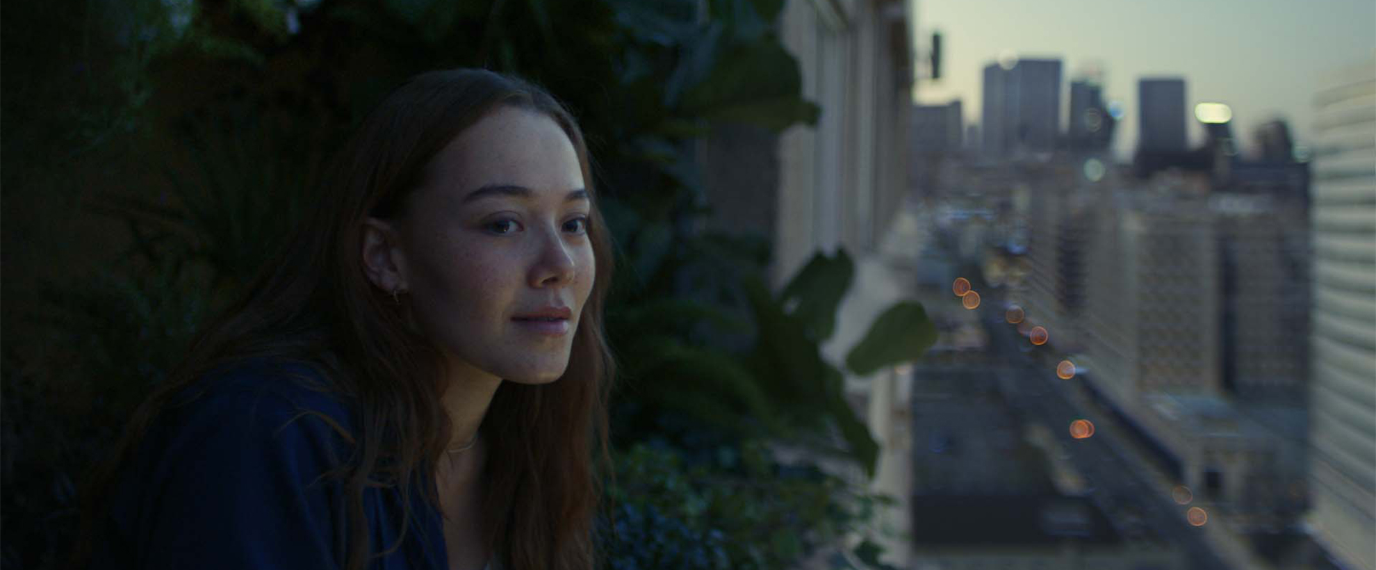 Concrete block of flats focusing on one balcony full of green plants whilst a woman looks out into the distance