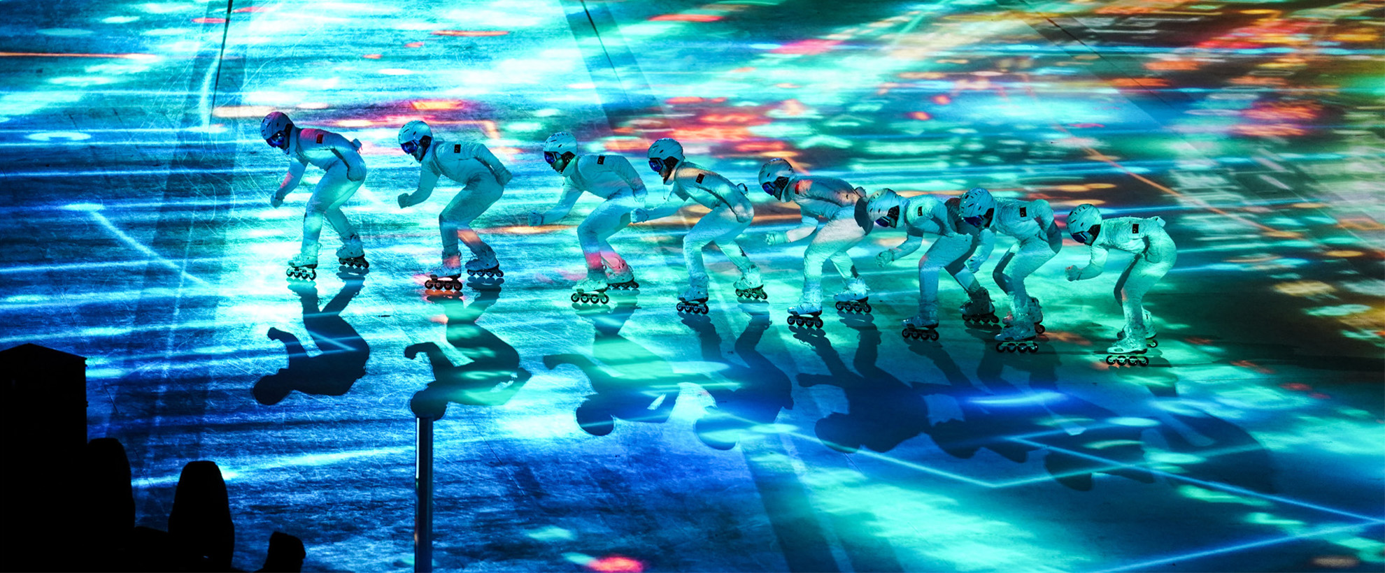Eight people wearing white snowsuits are ice skating in a line from right to left. There are blue and green lights projected on the ice. 