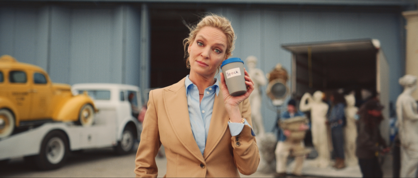 actress uma thurman raising eyebrows while holding a coffee cup that has 'spock' written on it in front of a film set