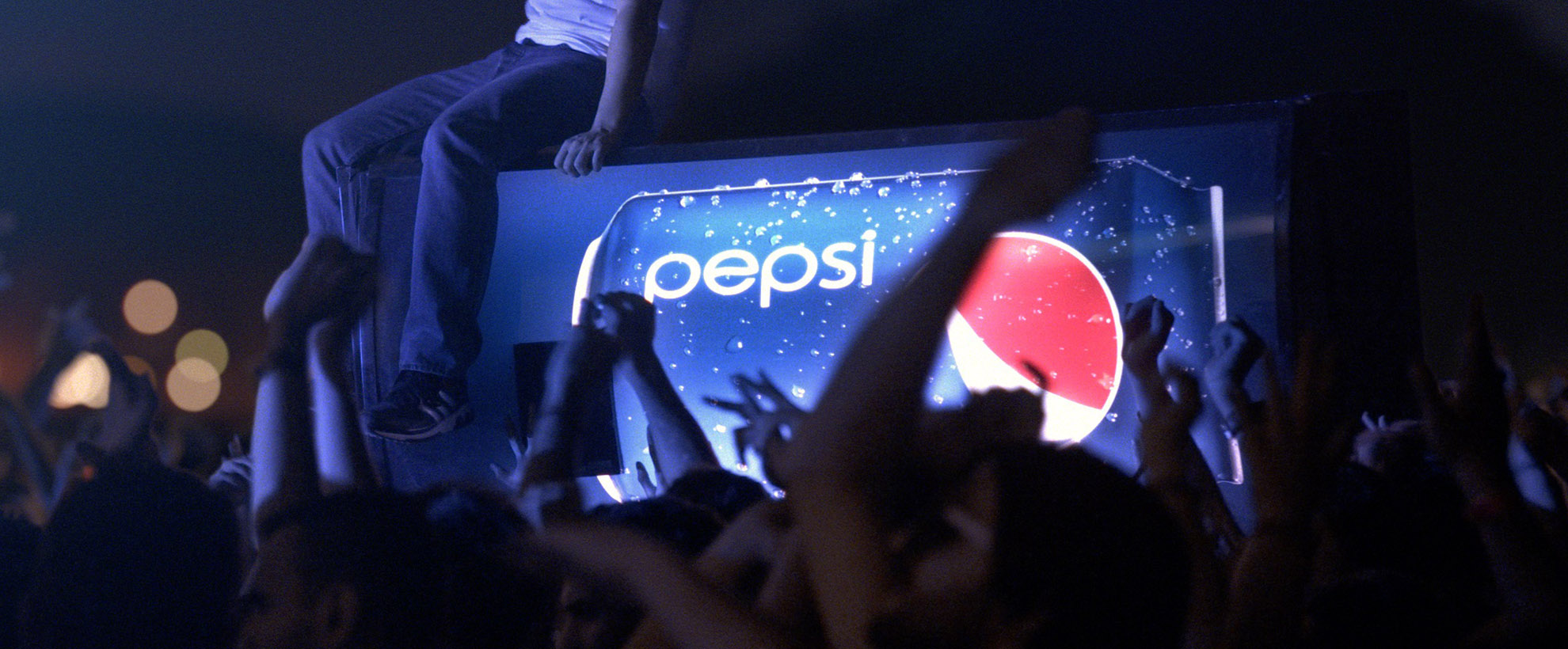 A Pepsi vending machine crowd surfs through a crowd, someone sits on top of the vending machine