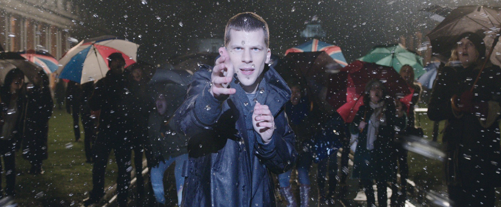 actor jesse eisenberg as j daniel atlas holding his hands in front of his chest as he is conjuring water droplets in the air. there are people with umbrellas walking by