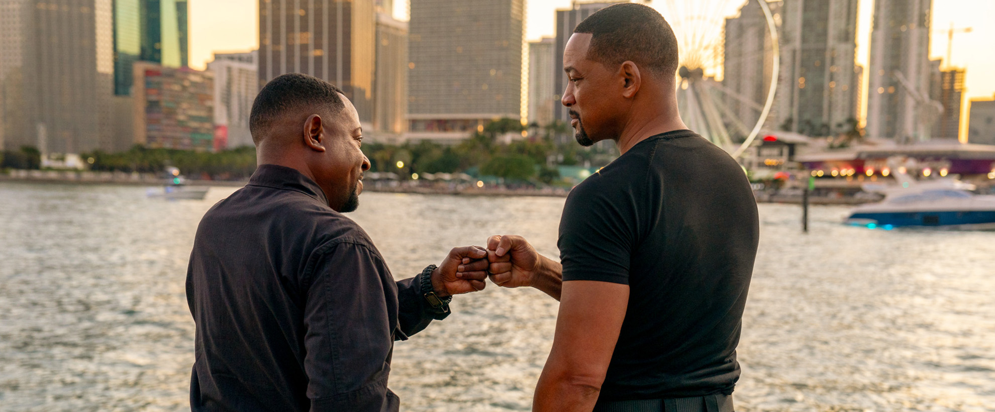 Two black men fist bumping in front of a body of water and a city skyline. 